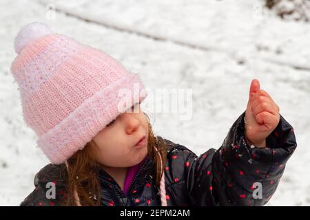 Porträt eines niedlichen, blauäugigen, braunhaarigen Mädchens, das einen rosa Hut und einen ablauen Mantel im Schnee trägt Stockfoto