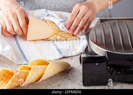 Eine kaukasische Frau rollt handgemachte Waffelkegel frisch aus Waffelkegel Hersteller Presse Maschine. Sie verwendet ein spezielles Rollwerkzeug auf dem Küchentuch. Einige i Stockfoto