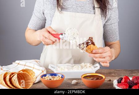 Eine Küchenchefin schaufelt hausgemachtes Vanilleeis auf handgemachte Waffel-Eiszapfen. Sie tauchte die Zapfen in Schokoladenschmelze und Walnussstücke f Stockfoto