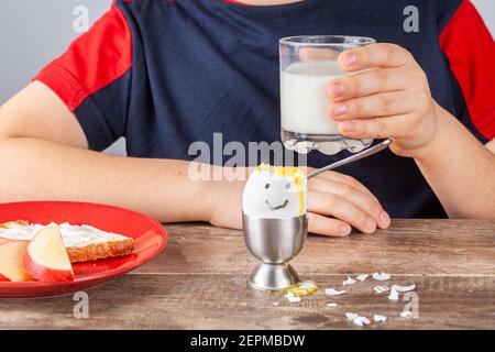 Frühstückstisch mit einfachen ausgewogenen nahrhaften Zutaten. Ein weich gekochtes Ei mit bemaltem Gesicht, eine Scheibe geröstetes Brot mit Frischkäse-Aufstrich Stockfoto