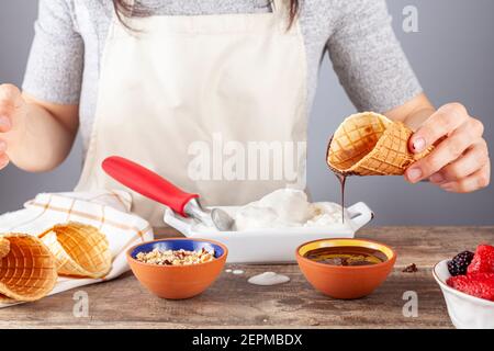 Eine Küchenchefin taucht hausgemachte Waffeleis-Zapfen in Schokoladenschmelze und Walnussstücke für einen handwerklichen Look. Andere handgerollte Kegel sind dort. A m Stockfoto