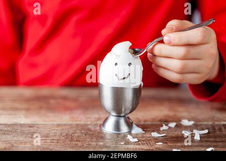 Ein kleines Kind isst weich gekochtes humpty dumpty Ei in Eierbecher auf einem hölzernen Frühstückstisch gelegt. Sie verwendet Löffel, um das Ei aus seiner Schale zu essen. Sie Stockfoto