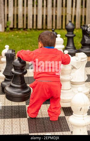 Ein kleiner Kleinkind Junge versucht, Schach im Hinterhof zu spielen. Es ist ein gigantisches Schachspiel mit einigen Stücken, die so groß sind wie er selbst. Er ist begeistert m Stockfoto