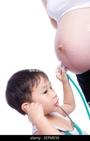 Kleiner asiatischer Junge in einem weißen Singlet, der den Bauch der schwangeren Mutter mit Stethoskop untersucht, Junge, der wie ein Arzt wirkt, isoliert auf weißem Hintergrund Stockfoto