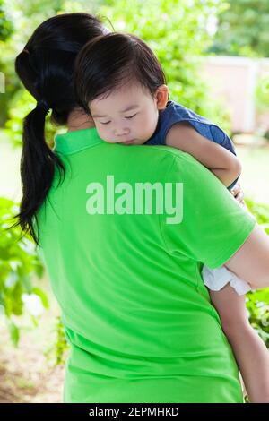 Mutter trägt ihre Tochter im Hinterhof über verschwommen grünen Natur Hintergrund, kleine asiatische Mädchen (thai) schlafen auf ihrer Mutter Schulter Stockfoto