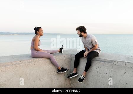 Fröhliche junge bärtige Sportler sprechen mit Inhalt ethnische weibliche Athletin Während er sich vor endlosem Meer ausruhte und sich ansah Stockfoto