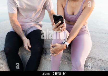 Junge froh Sportler mit ethnischen weiblichen Partner in Sportkleidung Während die Zeit zusammen mit dem Handy gegen Meer nach Schulung Stockfoto