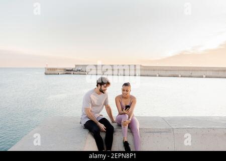 Junge froh Sportler mit ethnischen weiblichen Partner in Sportkleidung Während die Zeit zusammen mit dem Handy gegen Meer nach Schulung Stockfoto