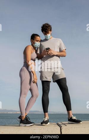 Nicht erkennbare ethnische Sportlerin in steriler Maske in der Nähe eines männlichen Partners, der auf dem Handy gegen das wellige Meer unter bewölktem Himmel im Internet surft Stockfoto