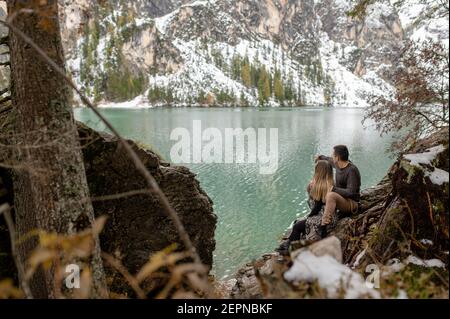 Voller Körper der liebevollen sanften Paar umarmt sich gegenseitig, während Sitzen auf Wald gegen Lago di Prags See umgeben von Verschneite Berge Stockfoto