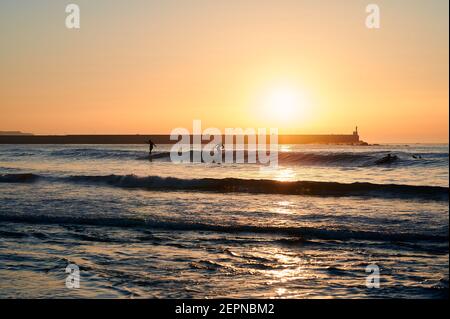 Silhouetten von Menschen schwimmen und surfen in Wellen des Meeres Unter strahlender Sonne, die am Himmel bei Sonnenuntergang scheint Stockfoto