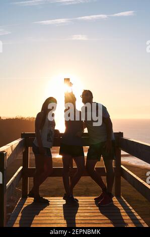 Lächelnde Touristen, die sich an Holzgeländer lehnen und sich selbst porträtieren Gegen Sonnenuntergang Himmel über ruhigen Meer Stockfoto