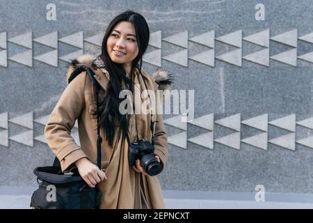 Positive asiatische Fotografin trägt einen warmen Mantel mit Fotokamera Steht gegen Steinwand mit geometrischem Dekor Stockfoto