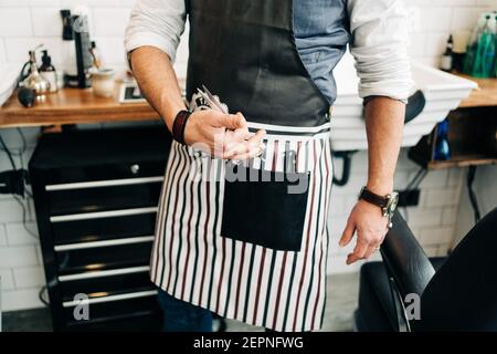 Crop unkenntlich männlichen Barbier mit Schere und Kamm in gestreift Schürze im Friseursalon Stockfoto