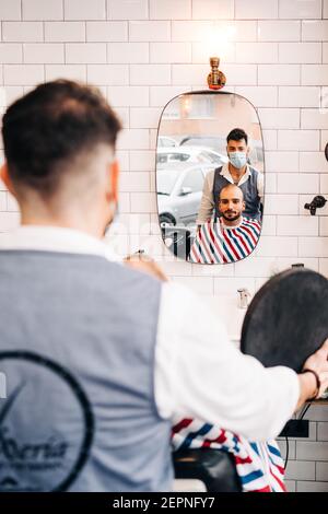 Rückansicht des anonymen männlichen Haarstylistin in der Maske nahe bärtig Kunde reflektiert im Spiegel im Friseurladen Stockfoto