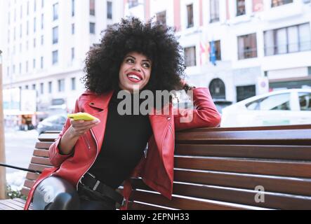 Stilvolle erstaunt afroamerikanische Frau Lesen Nachrichten auf Handy sitzen Auf der Bank in der Stadt Stockfoto