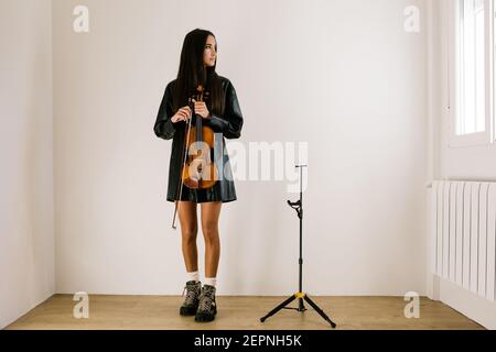 Ganzer Körper der Geigerin stehend mit Musikinstrument und Rücksichtsvoll auf das Fenster schauend Stockfoto