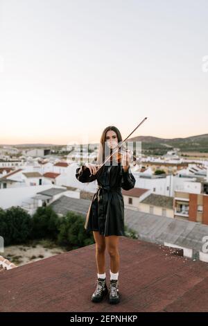 Junge schöne Musikerin in stilvollen Mini-Kleid hält akustische Violine und auf dem Dach in Wohnvorstadt stehen und suchen Weg auf sonnig Stockfoto