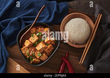 Nahaufnahme mala Tofu, chinesisches veganes Gericht, begleitet von einer Schüssel Reis auf einem mit Stoffen verzierten Holztisch Stockfoto