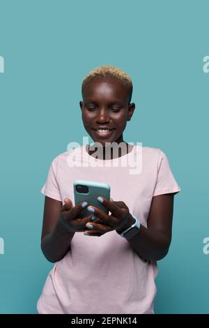 Ausdrucksstarke schöne afroamerikanische Frau mit kurzen Haaren und hell Maniküre Surfen auf dem Smartphone vor blauem Hintergrund Stockfoto