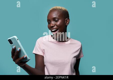 Ausdrucksstarke schöne afroamerikanische Frau mit kurzen Haaren und hell Maniküre Surfen auf dem Smartphone vor blauem Hintergrund Stockfoto