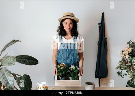 Junge Content Gärtnerin in Denim Overalls Blick auf die Kamera Mit Topfpflanzen mit blühenden Blumen in Holzkiste Stockfoto