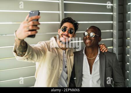 Glücklich bärtig ethnischen Mann mit Tattoo umarmt schwarzen Partner in Stilvoller Anzug und Sonnenbrille, während Sie sich selbst auf dem Handy porträtieren Stockfoto