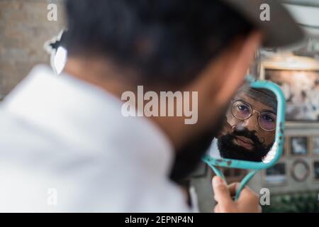 Rückansicht der Ernte bärtige Dandy ethnischen männlichen Friseur in Brillen spiegeln sich im Spiegel, während im Friseurladen der Schnurrbart geschwungen wird Stockfoto