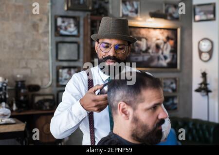 Stilvolle Dandy ernst ethnischen männlichen Friseur Trimmen Haare von Erwachsenen Kunde mit Elektroklipper im Friseursalon Stockfoto