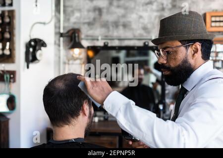 Stilvolle Dandy ernst ethnischen männlichen Friseur Trimmen Haare von Erwachsenen Kunde mit Elektroklipper im Friseursalon Stockfoto