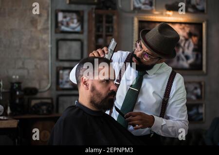 Stilvolle Dandy ernst ethnischen männlichen Friseur Trimmen Haare von Erwachsenen Kunde mit Elektroklipper im Friseursalon Stockfoto