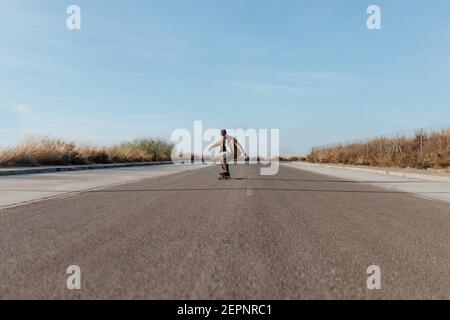 Ganzkörper junge bärtige männliche Skater in stilvollen tragen Reiten Skateboard entlang Asphaltstraße in der Landschaft Stockfoto