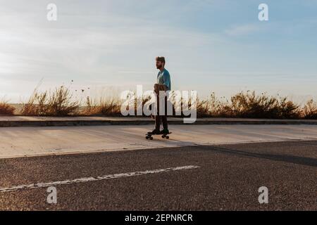 Ganzkörper junge bärtige männliche Reiten Skateboard mit Tasche und Jacke in der Hand entlang Bürgersteig in der Nähe Asphaltstraße Stockfoto