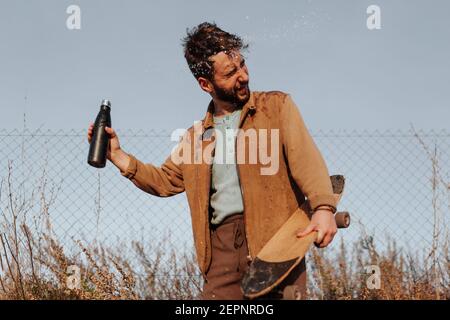 Junge bärtige männliche Skater mit geschlossenen Augen im lässigen Outfit Gießt Wasser aus der Flasche auf den Kopf, der in der Nähe des Zauns steht Auf dem Land Stockfoto