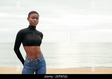 Junge sanfte afroamerikanische Frau in lässiger Kleidung zu suchen Kamera an der Meeresküste Stockfoto