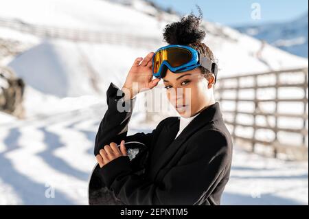 Hübsche afroamerikanische Athletin in modischer Kleidung und schützend Brillen stehen mit Snowboard Blick auf Kamera im Winter Stockfoto