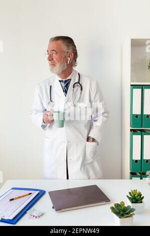 Leitender männlicher Arzt in Uniform mit Stethoskop mit Heißgetränk Im Krankenhaus bei Tageslicht wegschauen Stockfoto
