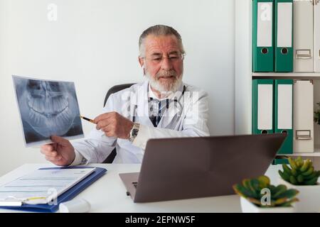 Älterer Kieferorthopäde mit Röntgenbild der Zähne während Video-Chat auf Netbook am Tisch im Krankenhaus Stockfoto