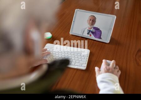 Von oben Rückansicht der Ernte anonyme ältere weibliche Patientin In TWS-Ohrhörern, die während des Videos mit dem Arzt auf einem Tablet sprechen Rufen Sie zu Hause an Stockfoto