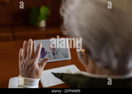 Von oben Rückansicht der Ernte anonyme ältere weibliche Patientin In TWS-Ohrhörern, die während des Videos mit dem Arzt auf einem Tablet sprechen Rufen Sie zu Hause an Stockfoto