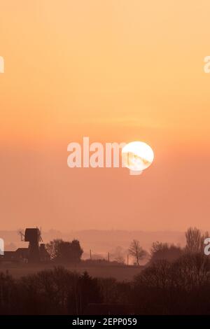 Sonnenaufgang über dem Norden der Landschaft von East Kent an einem nebligen Wintermorgen. Ehemalige Windmühle am Horizont mit der Sonne, eine große weiße Kugel, die über einer dünnen Wolkenschicht erscheint. Stockfoto