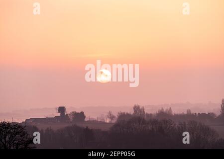 Sonnenaufgang über dem Norden der Landschaft von East Kent an einem nebligen Wintermorgen. Ehemalige Windmühle am Horizont mit der Sonne, eine große weiße Kugel, die über einer dünnen Wolkenschicht erscheint. Stockfoto