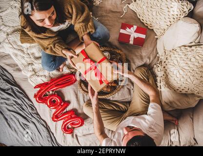 Junge glückliche Paar sitzt auf Decken im Bett zu Hause und Geschenke in Kisten über Obst und Zeichen Liebe auf dem Bett, Draufsicht. St. Valentinstag Konzept Stockfoto
