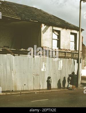 Eine Frau und zwei Kinder gehen an Häusern vorbei, die von Bomben in einer Belfaster Wohnsiedlung während der Unruhen im Jahr 1970s, Nordirland, Großbritannien, beschädigt wurden Stockfoto