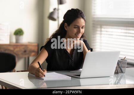 Lächelnde Frau, die auf den Laptop-Bildschirm schaut, Notizen macht, online studiert Stockfoto