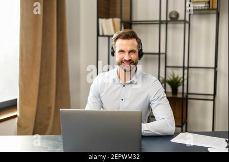 Lächelnder junger bärtiger Mann mit Headset und Blick auf die Kamera, positiver Hipster in formeller Kleidung, der in der Kundendienstabteilung arbeitet, Anrufe macht und entgegennimmt, im Heimbüro sitzt Stockfoto