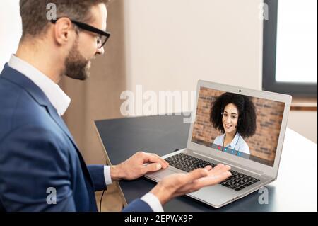 Ein intelligenter junger Büroangestellter in formeller Kleidung und Brille sitzt am Schreibtisch im Heimbüro, hat ein virtuelles Online-Meeting auf Laptop und spricht mit seiner Kollegin oder Freundin Stockfoto