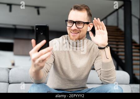 Gutaussehender, fröhlicher junger bärtiger Hipster in Brille mit schnurlosen Ohrhörern, der zu Hause auf einer Couch sitzt, auf den Bildschirm schaut, über ein Mobiltelefon einen Videoanruf abruft, winkt und hallo sagt Stockfoto