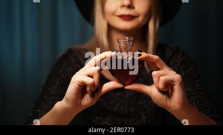 Zugeschnittenes Bild von Frau hält Herz geformten Glas der Liebe Trank. Schöne junge Frau in einem Hexenhut Stockfoto