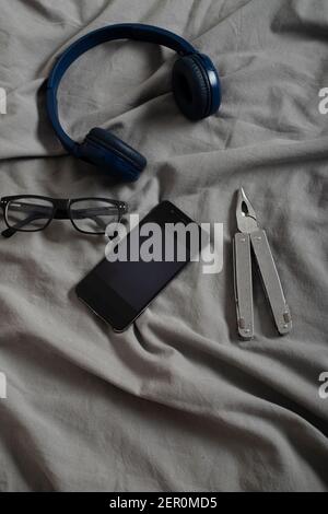 Mobiltelefon, Männerbrille und Mehrzweckgerät auf dem Bett mit grauen Laken Stockfoto